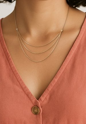 Collier délicat en or avec trois chaînes superposées porté autour du cou d'une femme, assorti à un chemisier rose saumon à col en V avec un seul bouton.