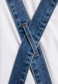 Denim hängslen korsar en vit tygbakgrund. Bandet är blått med en blekt struktur och gula sömmar, med en klassisk design.