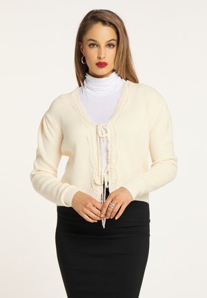 Cremefarvet cardigan med ribbet struktur, blondedetaljer langs udskæringen og snorelukning. Båret over en hvid højhalset trøje og sort nederdel.