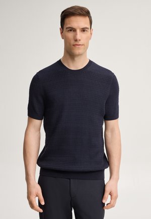 Homme debout devant un fond uni, portant un pull bleu marine à manches courtes texturé et un pantalon sombre, regardant droit devant lui.