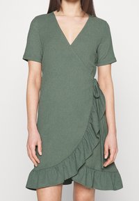 Vihreä kietaisumekko, jossa on lyhyet hihat, V-pääntie ja röyhelöyksityiskohtia helman varrella. Valmistettu teksturoidusta kankaasta. Ominaisuuksia ovat sivuriippu.