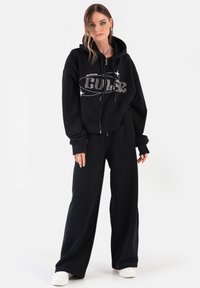 Czarna bluza z kapturem na zamek z logo "Cult" w srebrnym kolorze, zestawiona z luźnymi czarnymi spodniami dresowymi. Oba wykonane z miękkiego, wygodnego materiału. Białe trampki.