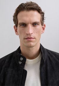 Mężczyzna z krótkimi, kręconymi brązowymi włosami, ubrany w czarną zapinaną kurtkę na białą koszulę, patrzy bezpośrednio w obiektyw na jednolitym tle.