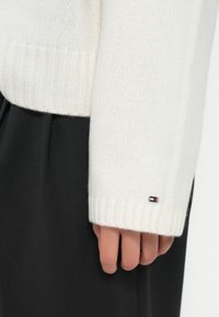 Suéter branco de malha com punhos canelados, apresentando uma pequena etiqueta de logotipo na manga. Combinado com calças escuras e texturizadas.