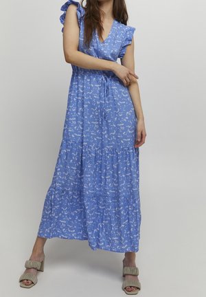 Femme posant dans une robe longue bleue à motifs floraux avec des manches volantées et des sandales à talons beige sur un fond uni.