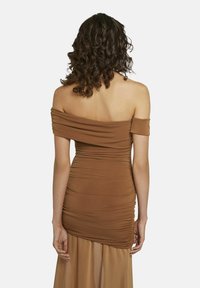 Vrouw met krullend donker haar, gekleed in een bruin, off-shoulder, gerimpeld jurkje met doorschijnende stof die onder de rok uitsteekt, met haar rug naar de kijker gekeerd.