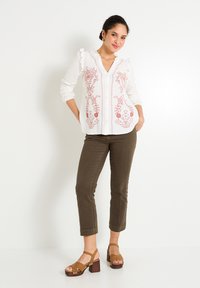 Blusa blanca con bordado floral en rojo, cuello con volantes y botones en la parte delantera, combinada con pantalones ajustados en verde olivo y sandalias de tacón cuadrado marrón.