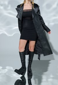 Svart läder trench coat över en åtsittande svart klänning, kombinerad med knähöga svarta plattformstövlar, vilket skapar en smidig och strukturerad silhuett.