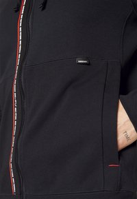 Sweat à capuche noir zippé avec logo Nike, poches latérales, coutures accent rouges, tissu texturé et finition douce.