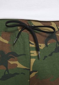 Kamouflage-byxor med gröna, bruna och gula mönster. Har en svart dragsko med metalliska spetsar. Mjuk bomullstyg.