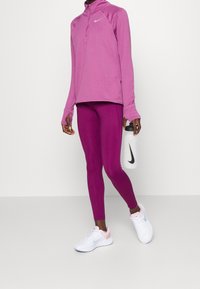 Personne portant des leggings de sport violets et un haut à manches longues, tenant une bouteille d'eau Nike blanche, et chaussée de chaussures de course Nike blanches et roses.