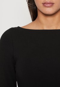 Musta vartalonmyötäinen paita venepääntiellä, valmistettu sileästä, joustavasta kankaasta. Pitkät hihat ilman näkyviä kuvioita tai metalliosia.