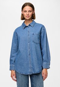 Modra denim srajca s gumbi in dolgimi rokavi, z ostrim ovratnikom in žepom na prsih. Tkanina ima mehak, rahlo teksturiran videz.