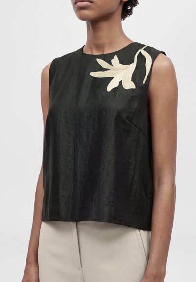 Mannequin portant un haut noir sans manches, orné d’une broderie florale couleur crème à l’épaule, et un pantalon beige clair.