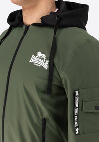 Olivovo zelená bunda na zips s čiernou kapucňou, obsahujúca biele logo, bočné vrecko na zips a štítok na rukáve. Hladká, vodeodolná tkanina.
