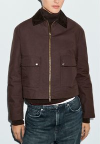 Braune, kurz geschnittene Jacke mit einem Reißverschluss vorne, zwei Fronttaschen und einem kontrastierenden schwarzen Cordkragen. Getragen über einem dunklen Rollkragenpullover und blauen Jeans.