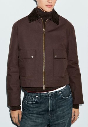 Giacca corta marrone con zip frontale, due tasche anteriori e colletto in cotone nero a contrasto. Indossata sopra un dolcevita scuro e jeans blu.