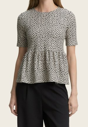 Žena v bílém krátkým rukávem peplum topu s černými nepravidelnými puntíky a volnými černými kalhotami na jednoduchém pozadí.