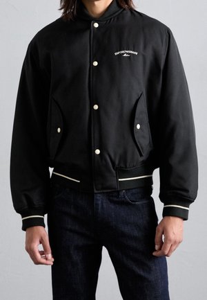 Mężczyzna w czarnej kurtce varsity z białymi napami i logo Emporio Armani na piersi, w połączeniu z ciemnoniebieskimi dżinsami.