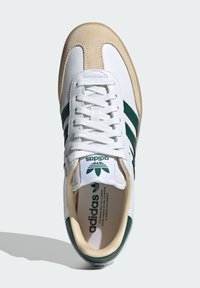 Sneaker in pelle bianca con righe verdi, cappuccio in camoscio beige, e lacci bianchi. Presenta un logo sulla linguetta e un interno imbottito.