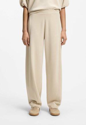 Pantalon beige droit taille haute avec une coupe ample, porté avec des baskets beige assorties et un chemisier beige clair.