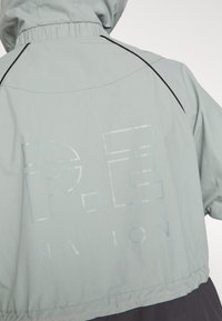 Chaqueta cortavientos de color gris claro con puños y dobladillo elásticos. Presenta un logotipo brillante de "P.E NATION" en la parte posterior, con detalles en negro.