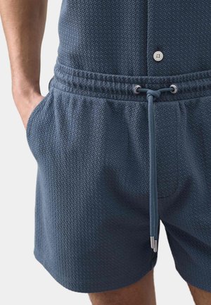 Uomo che indossa pantaloncini blu texturizzati con vita con coulisse e una mano in tasca, ritagliato in vita e cosce.