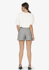 Cremefarbenes Baumwoll-T-Shirt mit kurzen Ärmeln, kombiniert mit grauen karierten Shorts, die große Gesäßtaschen haben. Das Modell trägt schwarze Sandalen.