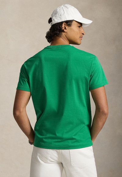 Žalias medvilninis trumpomis rankovėmis marškinėlis su apvalia iškirpte ir lygiu audiniu. Dėvimas su baltais kelnėmis ir balta kepure.