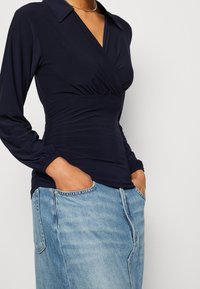 Marinblå långärmad topp med omlottdesign och elastiskt rynk vid midjan, kombinerad med en ljus tvättad jeanskjol med sidofickor.