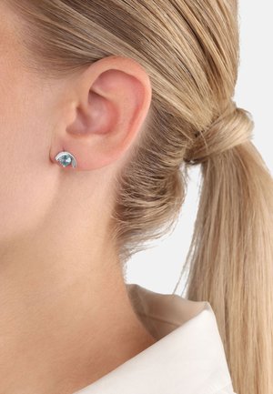 Orecchini in tonalità argento presentano un design curvo con una pietra preziosa blu chiaro al centro, su uno sfondo di pelle liscia.