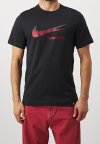 Musta puuvillainen t-paita, jossa on punainen ja harmaa Nike Swoosh -logo sekä punaisella painettu "LIVERPOOL FC" -teksti. Lyhyet hihat, klassinen istuvuus.