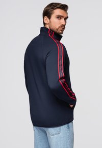 Marineblaue Zip-Jacke mit hohem Kragen, roten Streifenakzenten an den Ärmeln und entspannter Passform. Hergestellt aus weichem Material, geeignet für Freizeitkleidung.