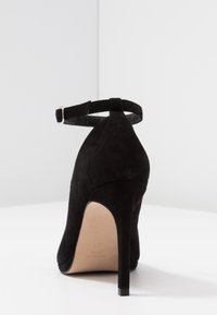 Svart mocka högklackad sko med stängd bak och justerbar vristrem. Slät textur och spetsig tådesign.