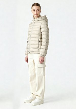 Femme debout portant une veste matelassée à capuche beige clair, un pantalon cargo crème et des baskets plateforme blanches, sur un fond uni.