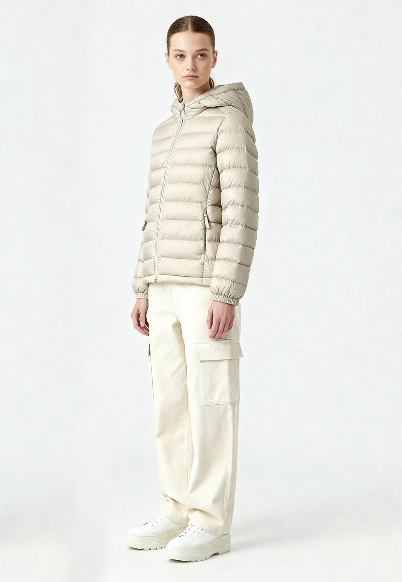 Femme debout portant une veste matelassée à capuche beige clair, un pantalon cargo crème et des baskets plateforme blanches, sur un fond uni.