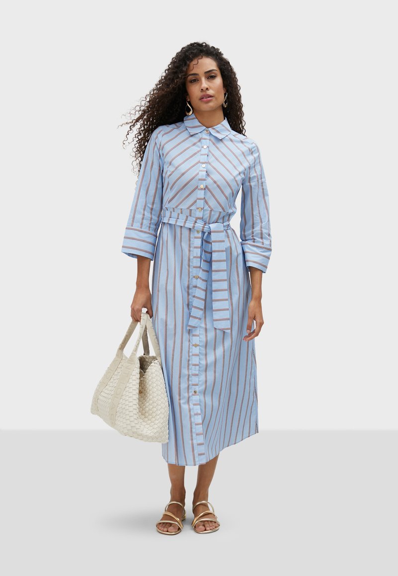 Light blue, striped mid-length shirt dress with a tied waist, button-down front, and three-quarter sleeves. Paired with a woven bag.