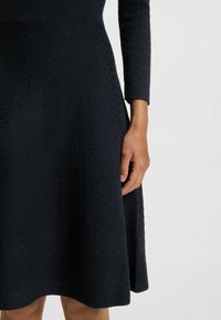 Robe navy à manches longues, en tissu texturé avec un motif subtil, et une jupe évasée. Gros plan sur la taille et la position de la main.