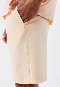 Pantaloni corti realizzati in tessuto pesca chiaro con una texture liscia, dotati di una tasca laterale e di una linea dritta. L'orlo è rifinito con cura.