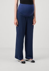 Pantalons de maternité navy avec une taille texturée et extensible. Coupe ample avec des jambes larges et un tissu froissé. Associés à des ballerines argentées et noires.