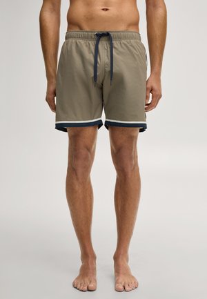 Homme portant un short de bain beige avec une bordure bleu marine et blanche, debout pieds nus sur un fond clair uni.