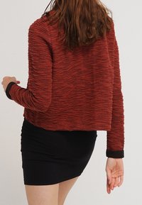 Rød tekstureret strikket cropped jakke med lange ærmer og sorte manchetter, parret med en tætsiddende sort nederdel.