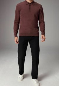 Burgundy long-sleeve polo sweater with a collar, three-button placket, worn with black pants and white sneakers, against a plain gray background.