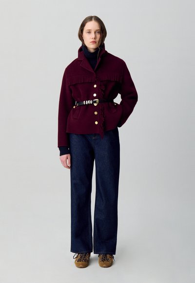 Chaqueta de lana burdeos con detalles de flecos, botones dorados, cintura ceñida con cinturón, combinada con jeans oscuros de pierna ancha y zapatos marrones con cordones. Fondo liso.