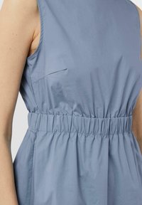 Helles blaues Stoffkleid mit gerafftem Taillenbund, ärmellosem Design und einer kleinen Tasche auf der linken Seite für zusätzliche Details.