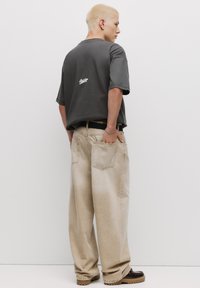 Grå kortærmet skjorte med logo på ryggen, parret med løse beige jeans med to baglommer og en bleget tekstur. Brune sko.
