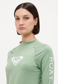Lys grønn langermet rash guard med en hvit logo på brystet og tekst som sier "ROXY" langs ermet. Glatt, tett passform.