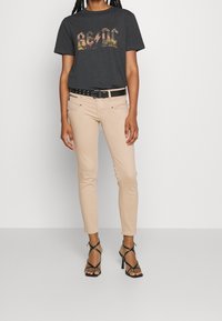 T-shirt gris avec un graphisme AC/DC, jean skinny beige clair, ceinture noire avec des clous en argent, et sandales à talons hauts noires à lanières.