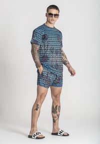 Modrá a černá vzorovaná sada trička s krátkým rukávem a šortek s opakujícím se textem. Model nosí černé sluneční brýle a bílé sandály s textem.