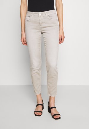 Jeans beige clair avec une coupe slim, cinq poches et une fermeture éclair. Associés à des sandales à talons noires. Texture douce et délavée.